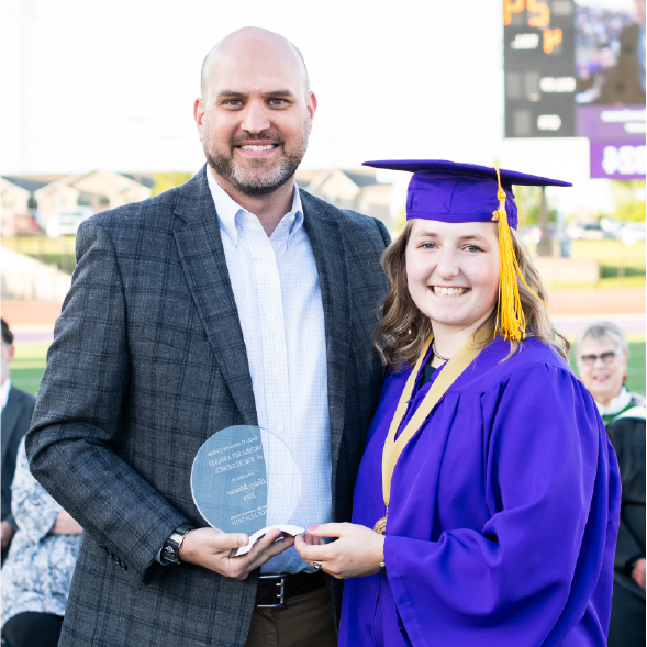 Female graduate in purple gown receives award from man in gray suit jacket.